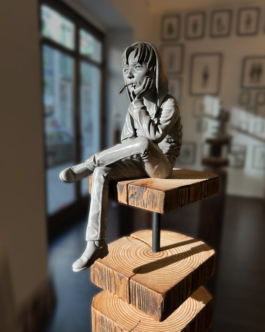 Skulptur einer sitzenden Figur auf einem Holzblock, umgeben von Kunstwerken an der Wand.