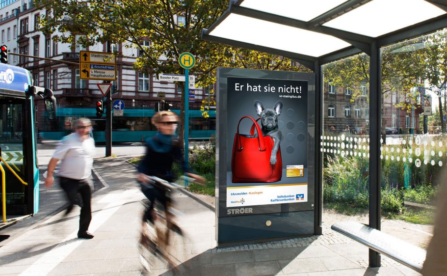 Citylight Wartebereich mit Werbung für eine rote Tasche, neben Radfahrern und Passanten.