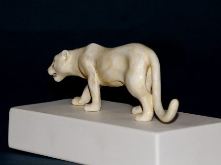 Elfenbeinfigur einer Katze in seitlicher Ansicht auf einem Sockel.